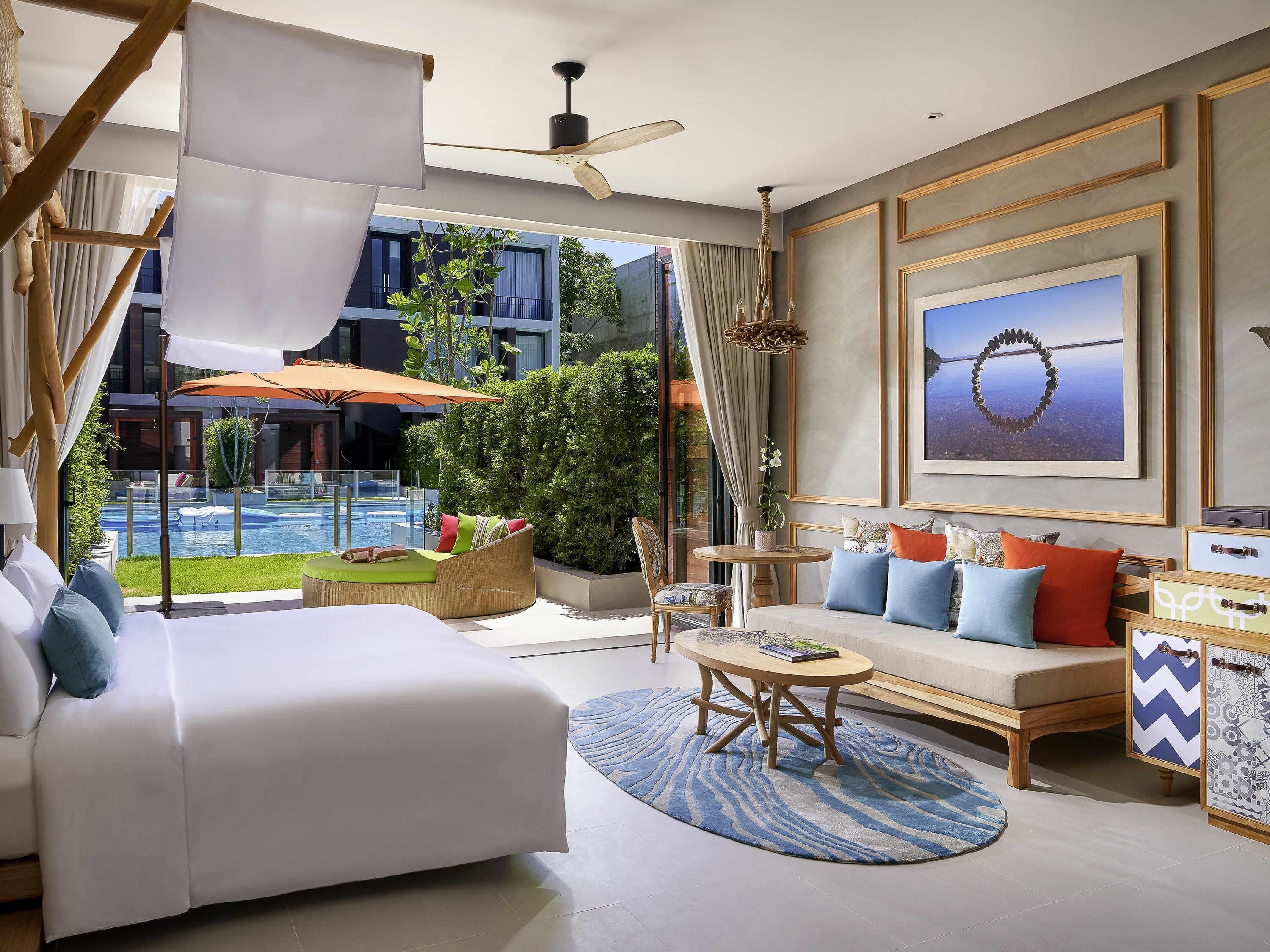 A modern hotel room with a large bed, a sofa, a round coffee table, and a view of a swimming pool.