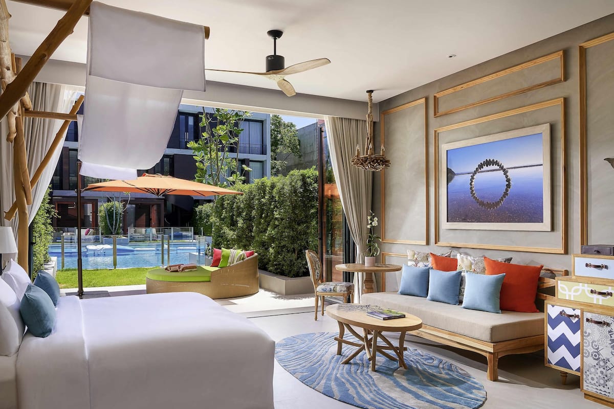 A modern hotel room with a large bed, a sofa, a round coffee table, and a view of a swimming pool.