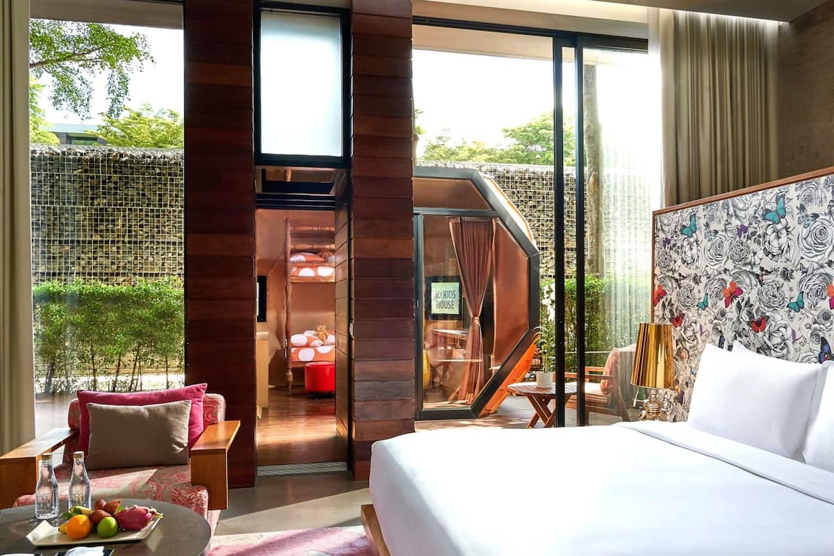 Hotelkamer met een bed, een eettafel met fruit, een bank en uitzicht op een tuin en een gebouw.