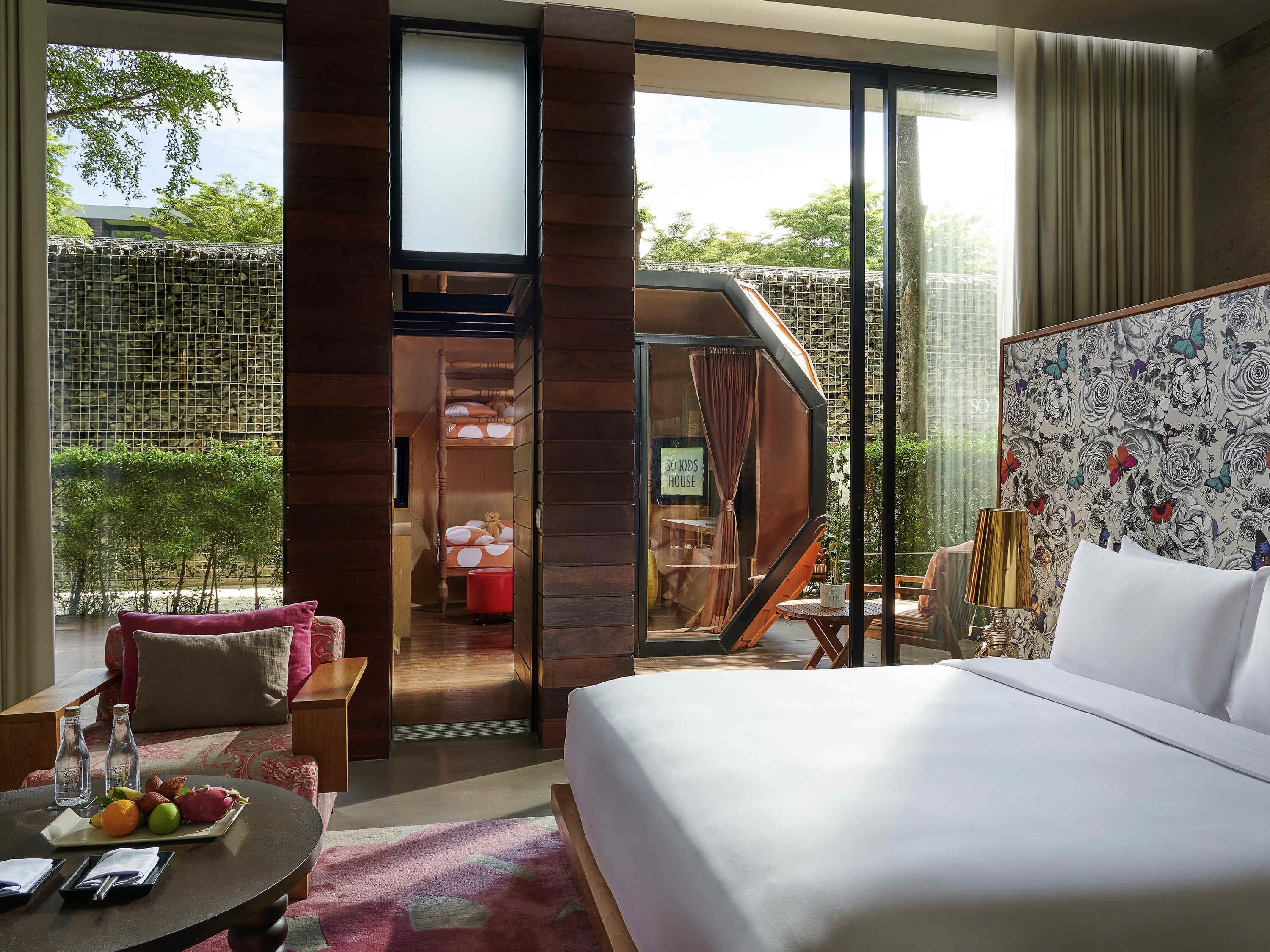 A hotel room with a bed, a dining table with fruit, a sofa, a balcony with a view of trees, and a children's play area.
