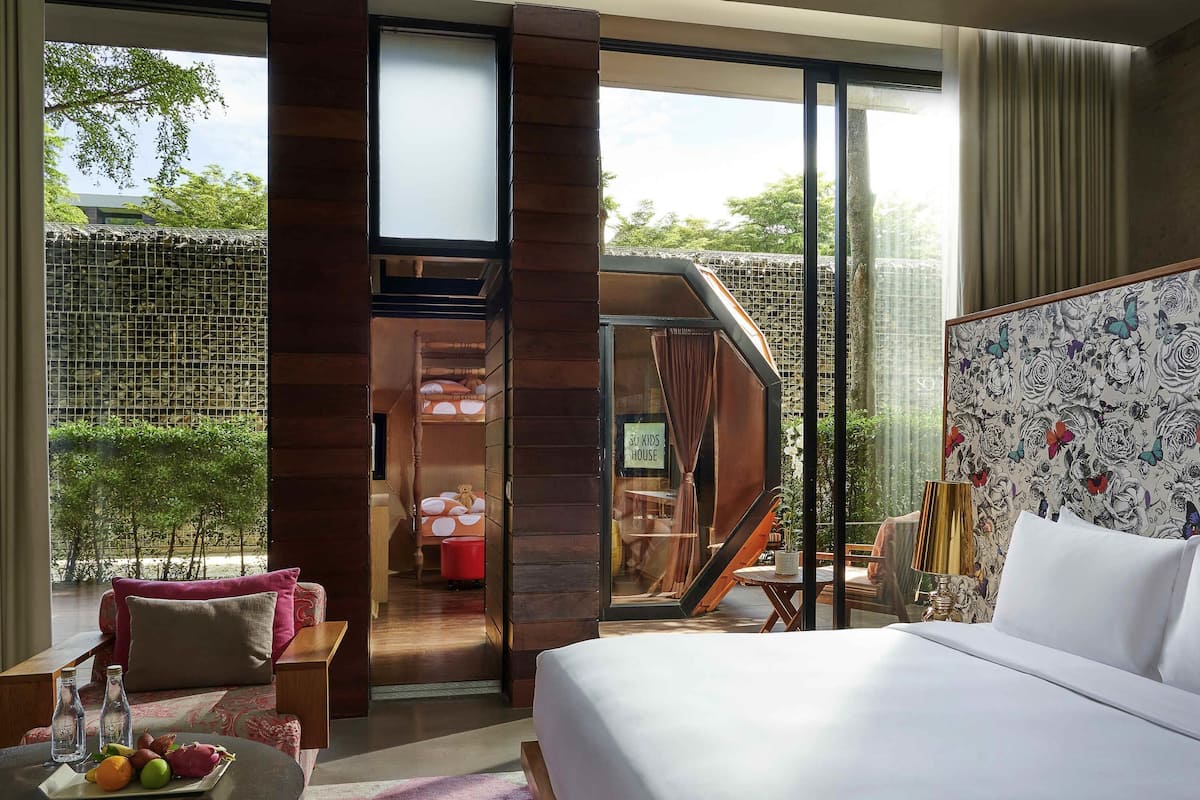 A hotel room with a bed, a dining table with fruit, a sofa, a balcony with a view of trees, and a children's play area.