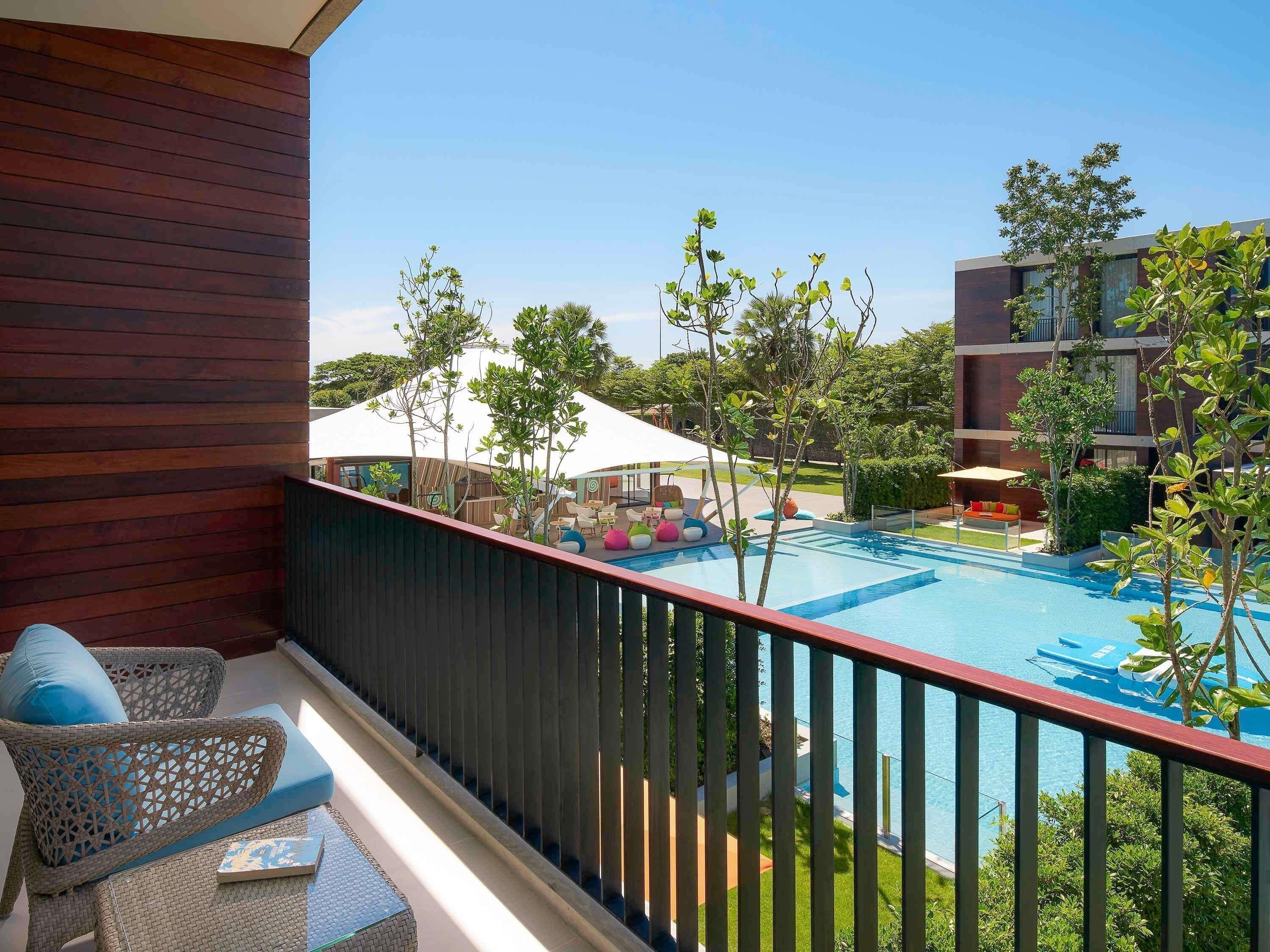 Un balcón con mobiliario de mimbre, una piscina y vistas a un edificio con toldo blanco.