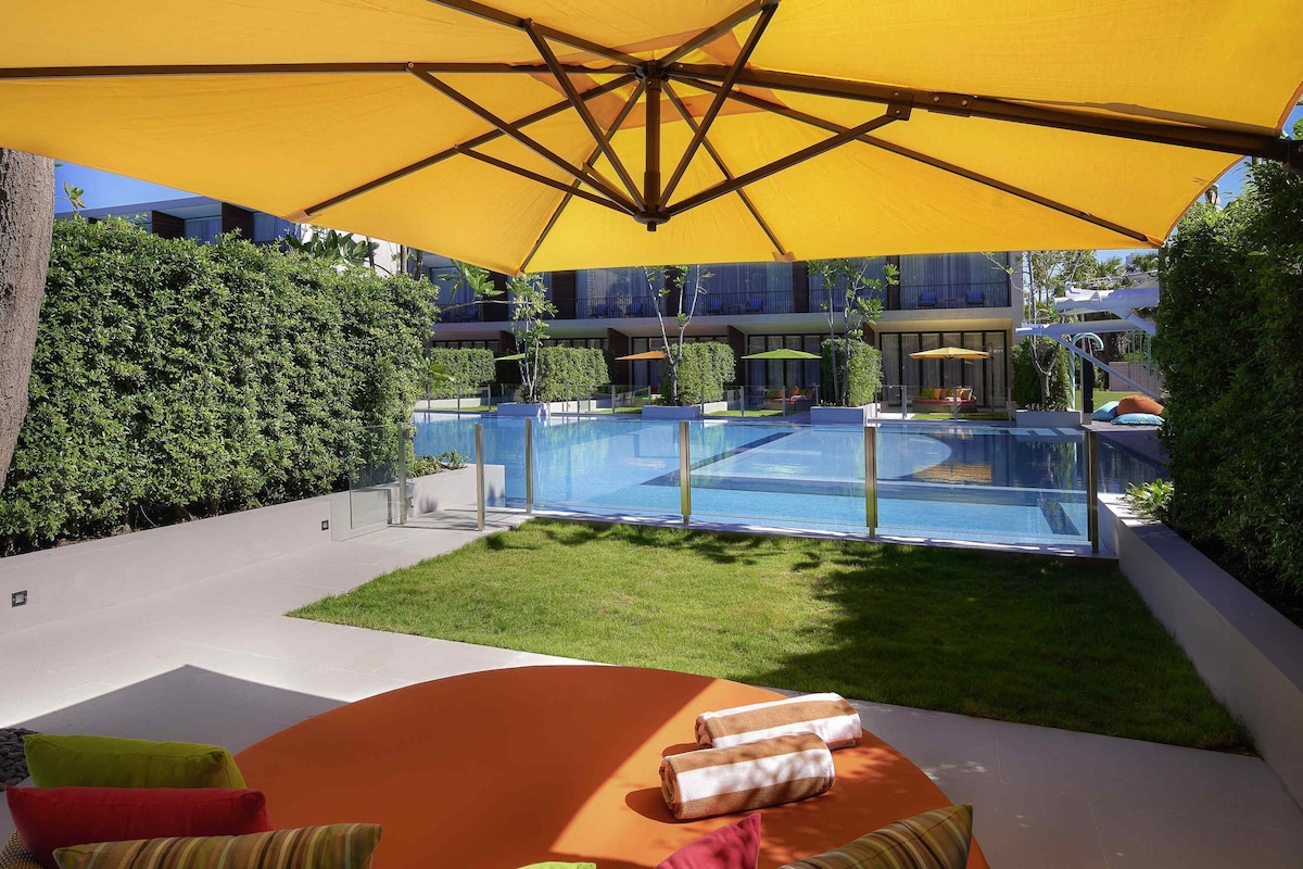 Zona de piscina con una sombrilla amarilla, mobiliario exterior y vistas a un edificio con piscina.