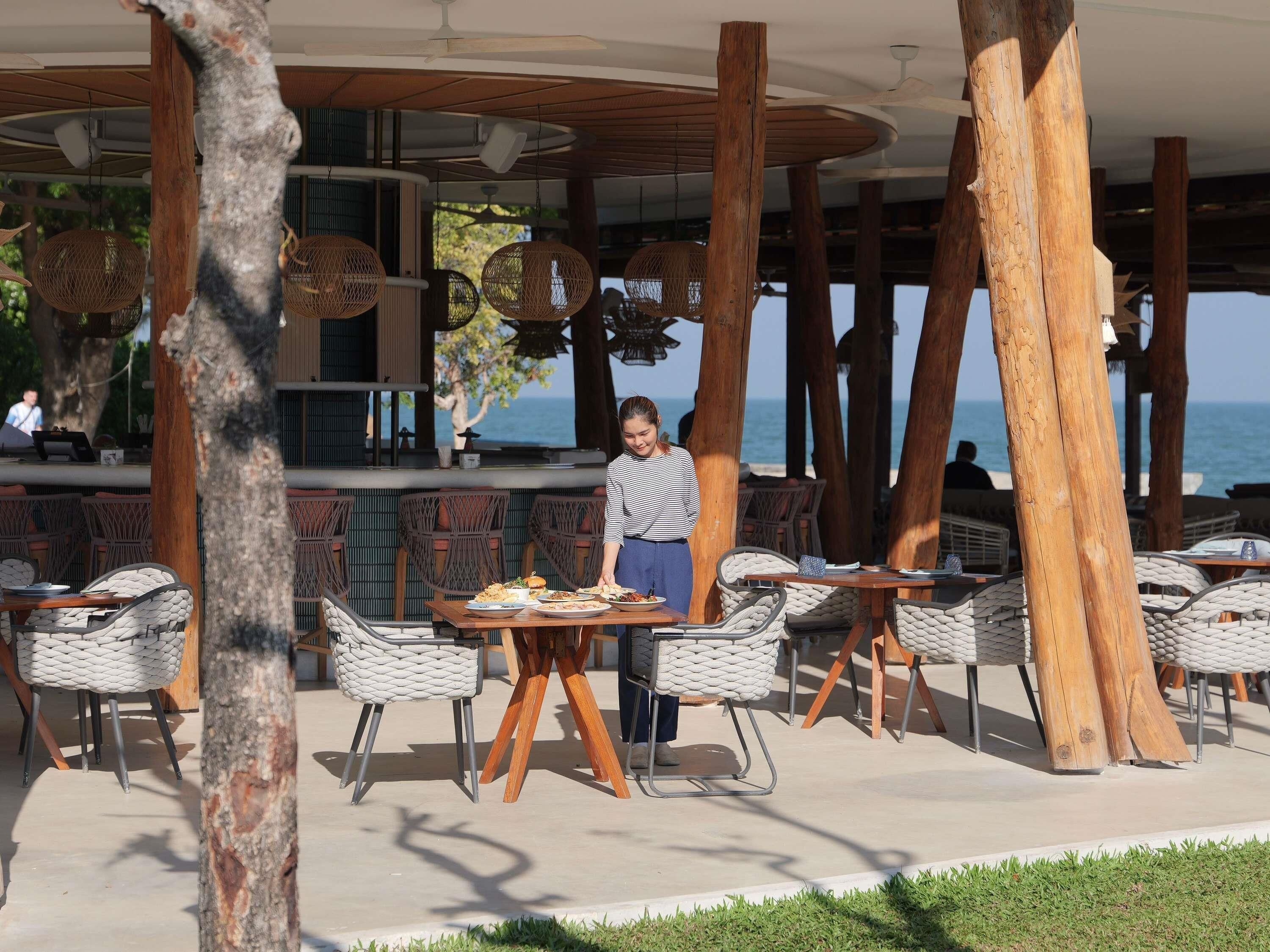 An outdoor dining area with wicker chairs and tables, wooden pillars, and hanging rattan lamps.