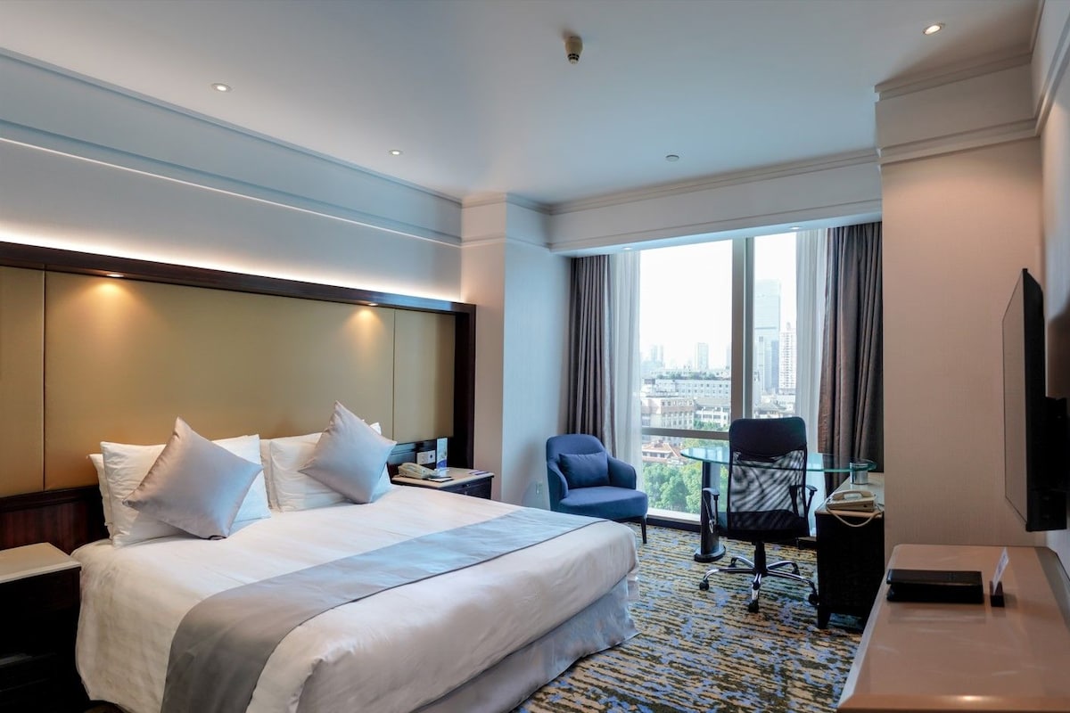Ein Hotelzimmer mit einem großen Bett, einem Schreibtisch mit Stuhl, einem Fernseher und Blick auf die Stadt durch große Fenster.