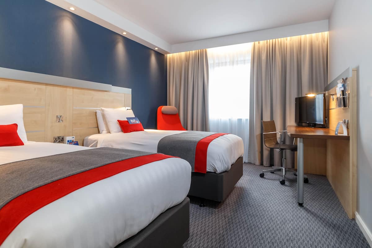 Habitación de hotel con dos camas, un escritorio, una silla, un televisor y un ventanal con cortinas.