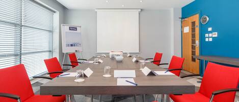 A conference room with a long table, red chairs, a whiteboard, and a projector screen.