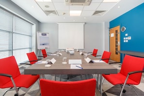 A conference room with a long table, red chairs, a whiteboard, and a projector screen.