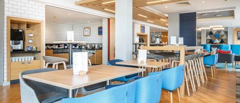 A modern cafe with wooden tables and blue chairs, a coffee machine, and a menu board.