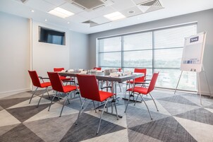 Un salón de conferencias con una gran mesa, sillas rojas, un atril blanco para rotafolio y un amplio ventanal con persianas.