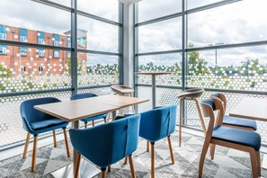 Un espacio interior moderno con mesas y sillas, ventanas amplias y vistas a edificios y áreas verdes.