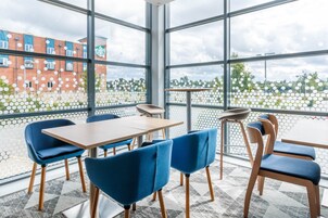 Un espacio interior moderno con mesas y sillas, ventanas amplias y vistas a edificios y áreas verdes.