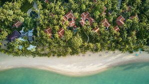 Vista aérea de un resort con varias construcciones tipo bungalow rodeadas de palmeras, una playa de arena y aguas turquesas cristalinas.