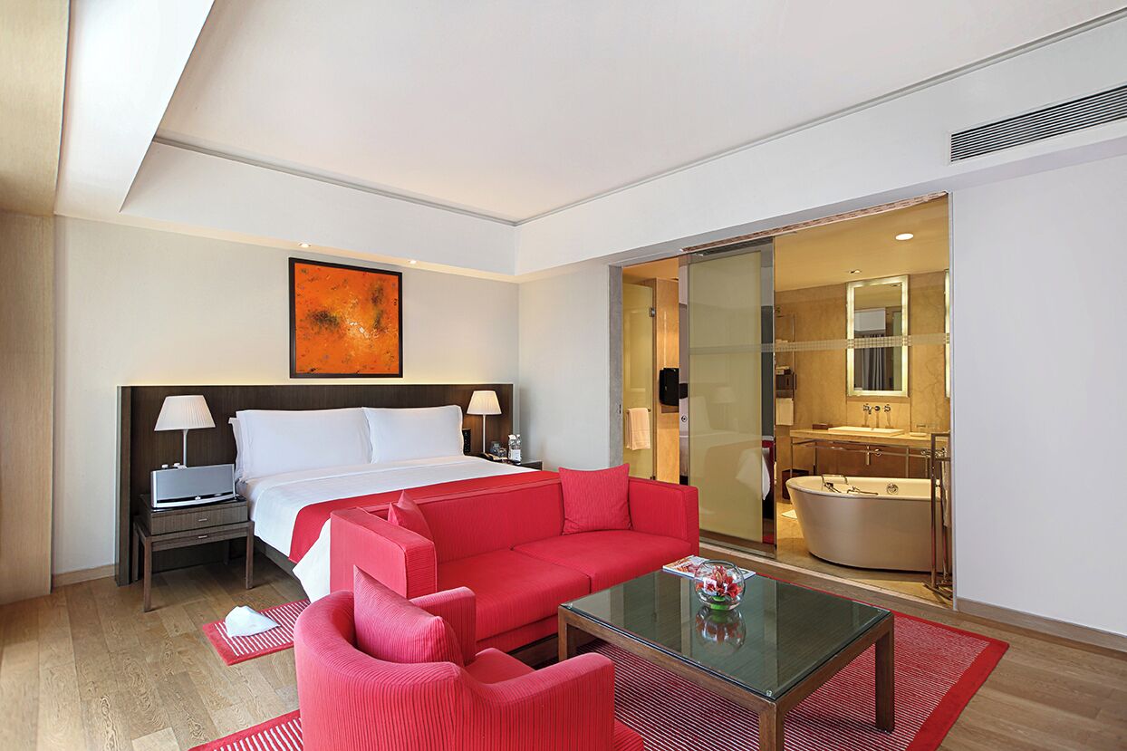 A hotel room with a bed, a red sofa, a glass coffee table, a nightstand, and a bathroom visible through a glass door.