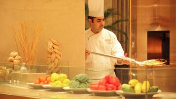 A chef in a white uniform holding a large serving spoon, standing behind a buffet counter with various food items like breadsticks, pastries, and fruits.