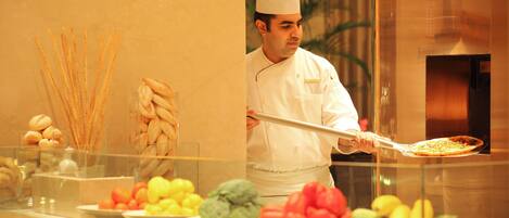 A chef in a white uniform holding a large serving spoon, standing behind a buffet counter with various food items like breadsticks, pastries, and fruits.