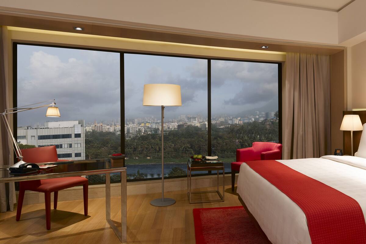 A hotel room with a large bed, a desk with a chair, a red chair, a lamp, and a view of the city.
