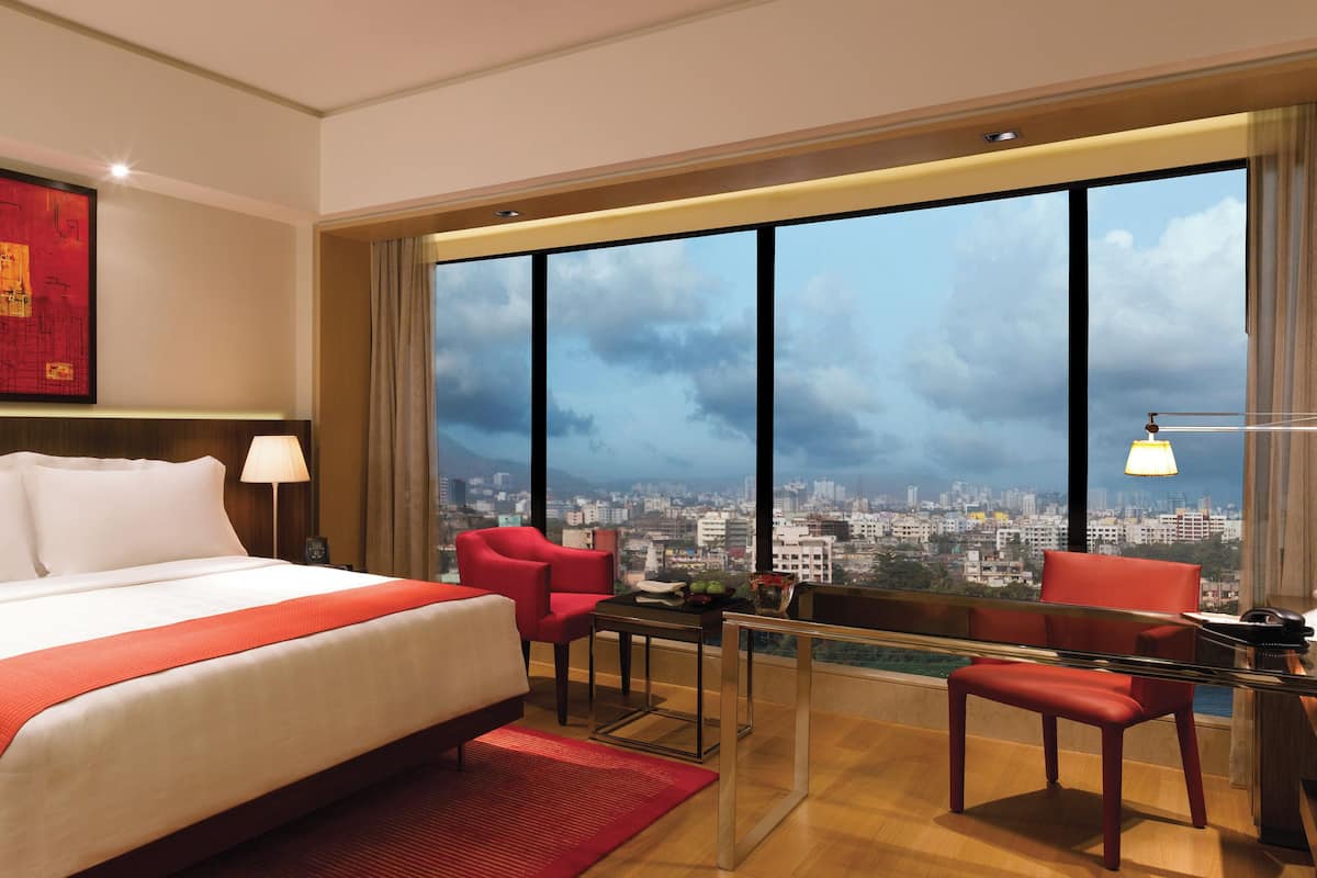 A hotel room with a large bed, a desk, two red chairs, a city view, and a red artwork on the wall.