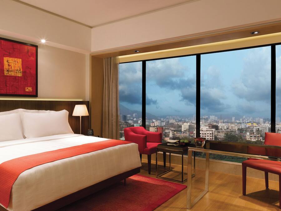 A hotel room with a large bed, a desk, two red chairs, a city view, and a red artwork on the wall.