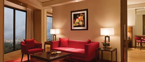 A modern hotel room with a red sofa, two side tables with lamps, a coffee table, and a large window offering a city view.