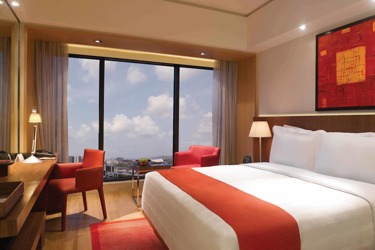 A hotel room with a large bed, a desk with a lamp, two chairs, a view of the city through large windows, and a red abstract painting on the wall.