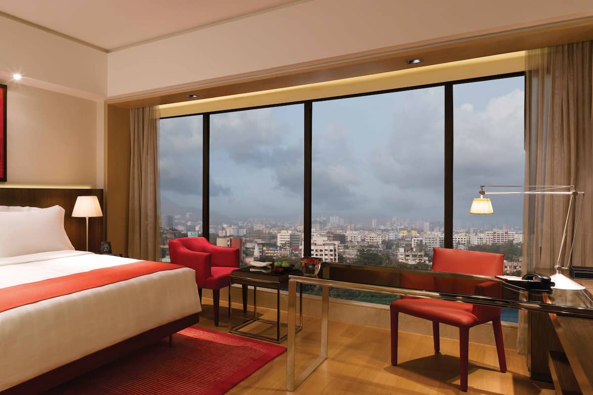 A hotel room with a large bed, a desk with two chairs, a city view, and a red artwork on the wall.