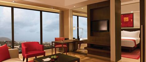 A modern hotel room with a large window, a flat-screen TV, a desk with a lamp, a bed, and red chairs.