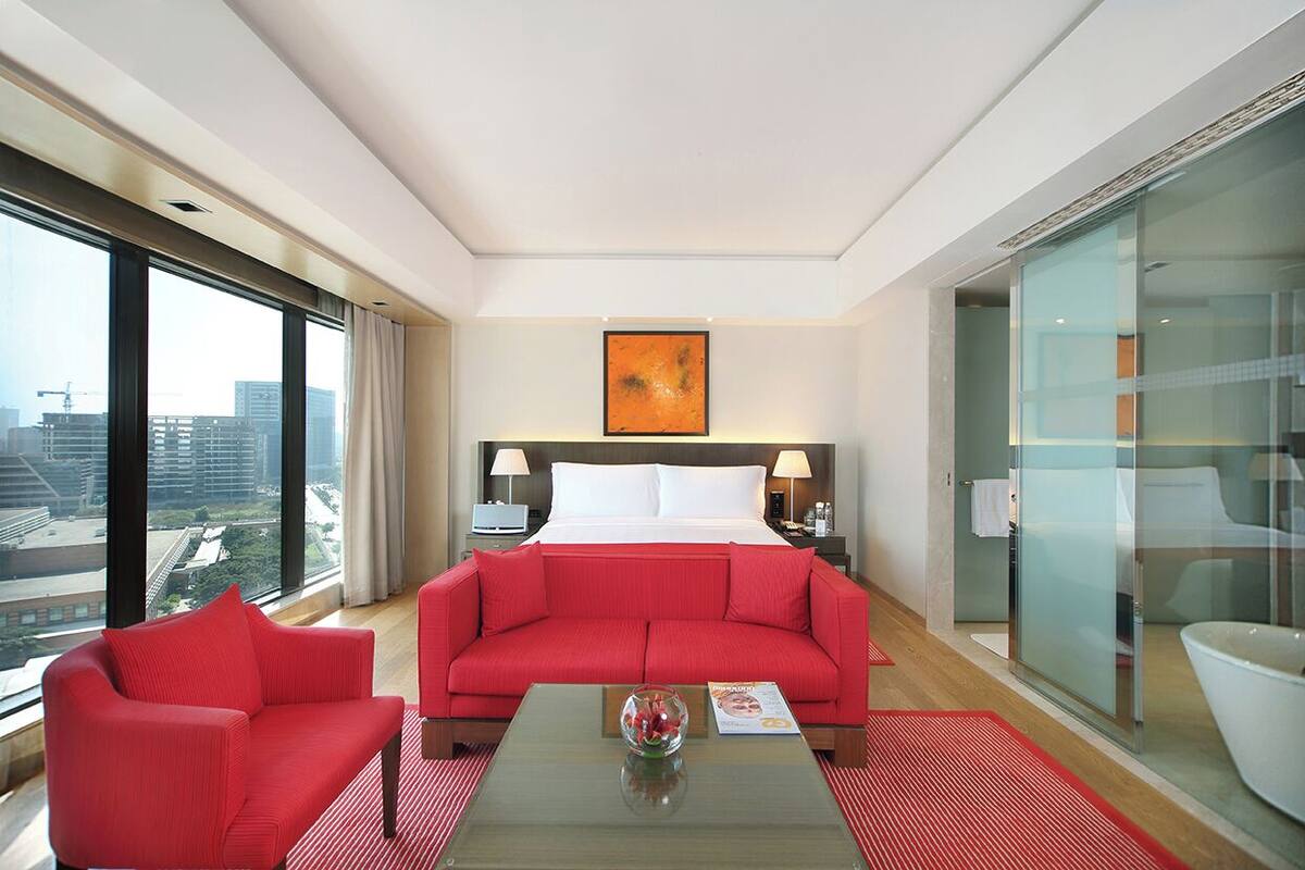 A hotel room with a large bed, a red sofa, a glass-enclosed bathroom, and a city view.