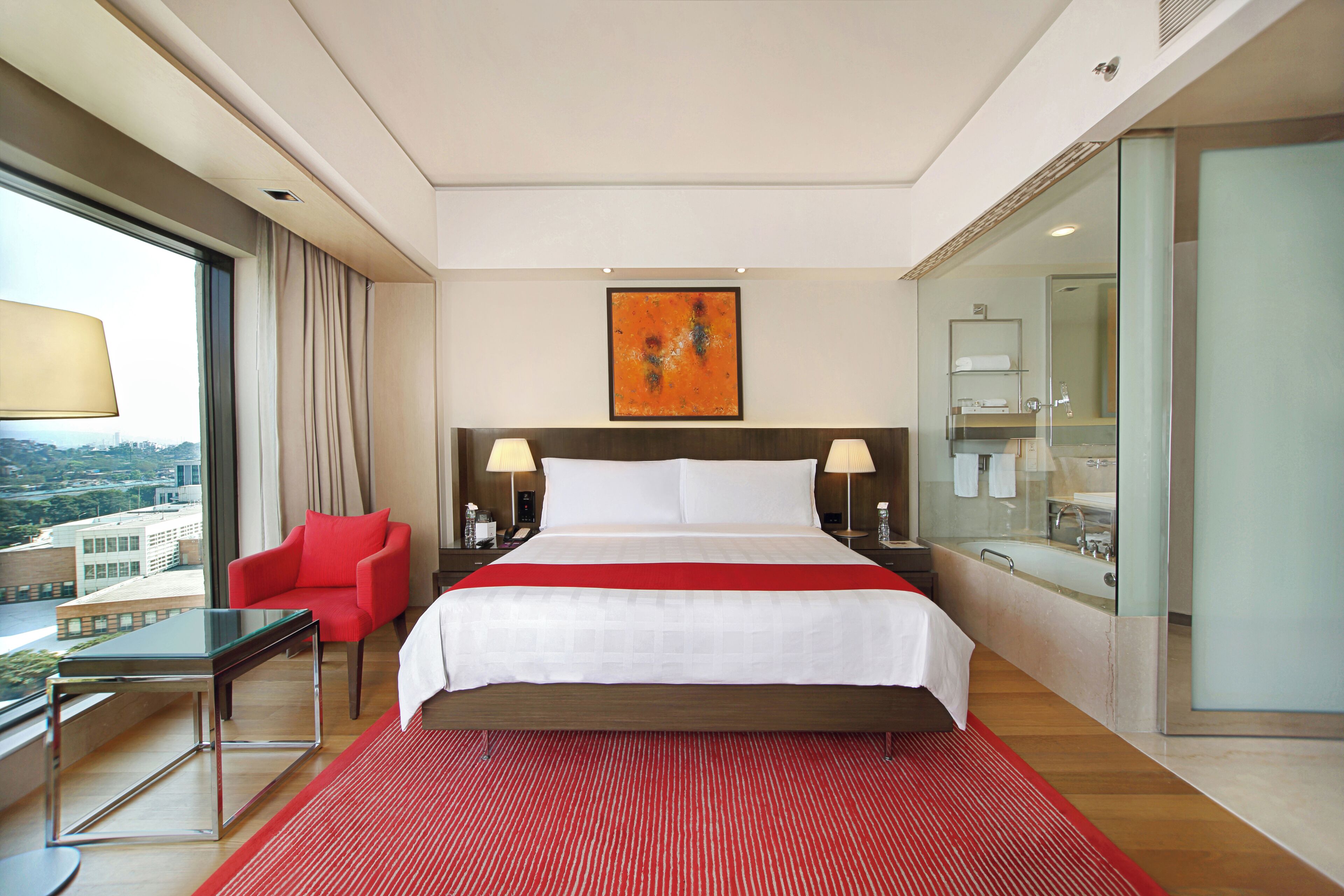 Camera d'albergo con un letto grande, una poltrona rossa, un tavolino in vetro, un tappeto a righe rosse e bianche, vista sulla città e un bagno moderno con doccia in vetro.