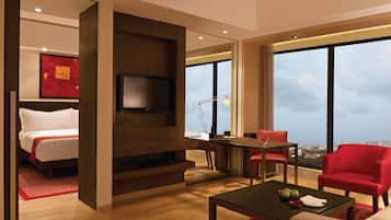 Una habitación de hotel moderna con cama, televisor montado en un panel de madera, escritorio con lámpara, una silla roja y vistas de la ciudad a través de amplias ventanas.