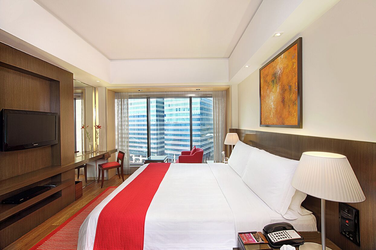 A hotel room with a large bed, a TV, a desk with a lamp, and a view of tall buildings.