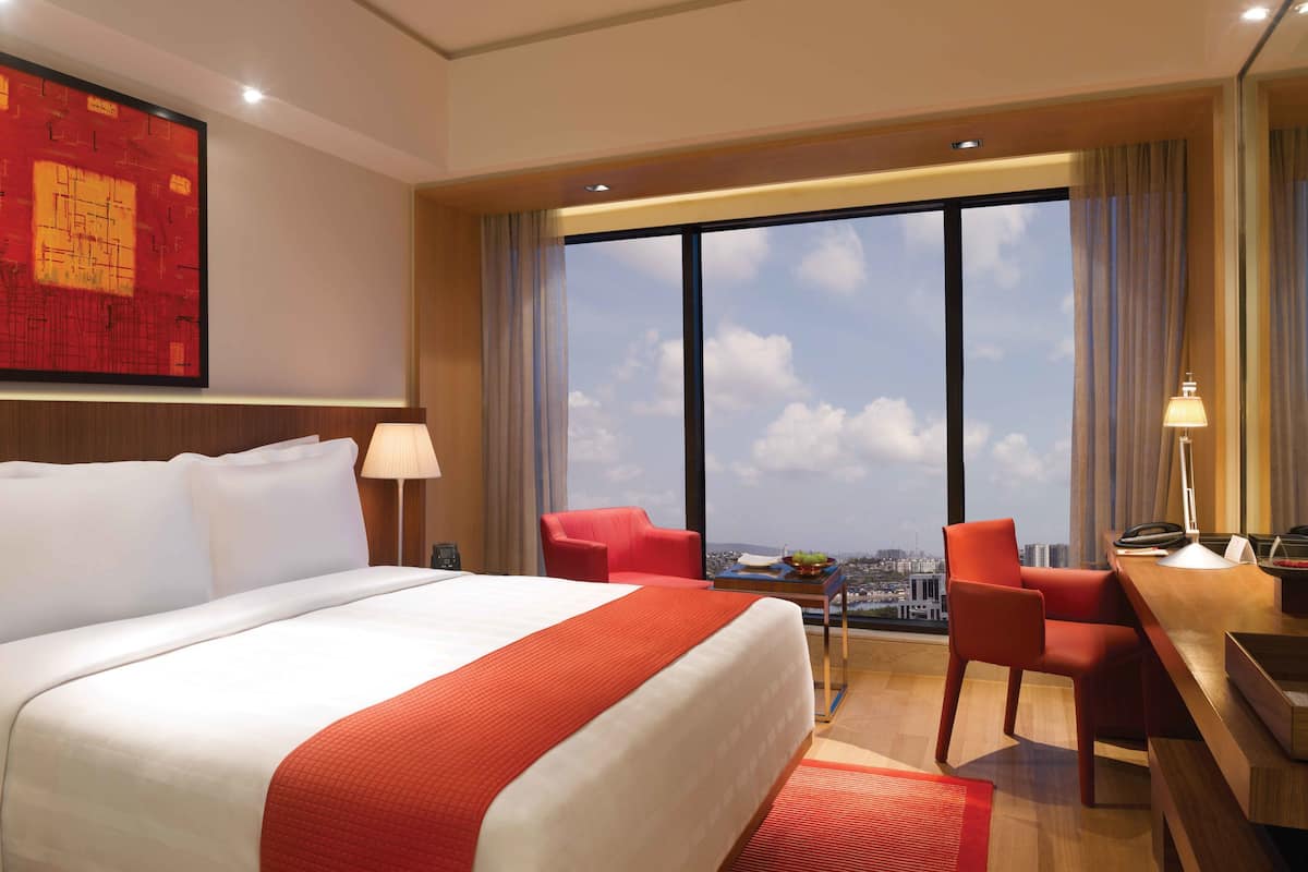 A hotel room with a large bed, a desk with a chair, a desk lamp, a telephone, and a view of the city through large windows.