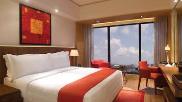A hotel room with a large bed, a desk with a chair, a desk lamp, a telephone, and a view of the city through large windows.