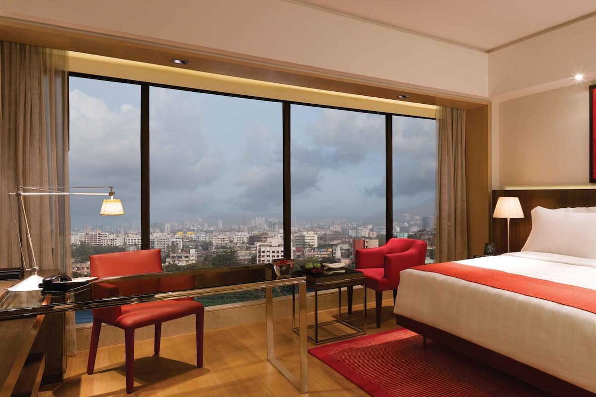 A hotel room with a large bed, a desk with a chair, a red chair, a view of the city through large windows, and a red artwork on the wall.