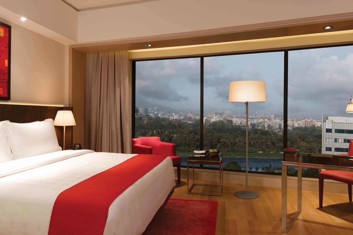 A hotel room with a large bed, a red armchair, a desk with a lamp, and a view of a cityscape through large windows.