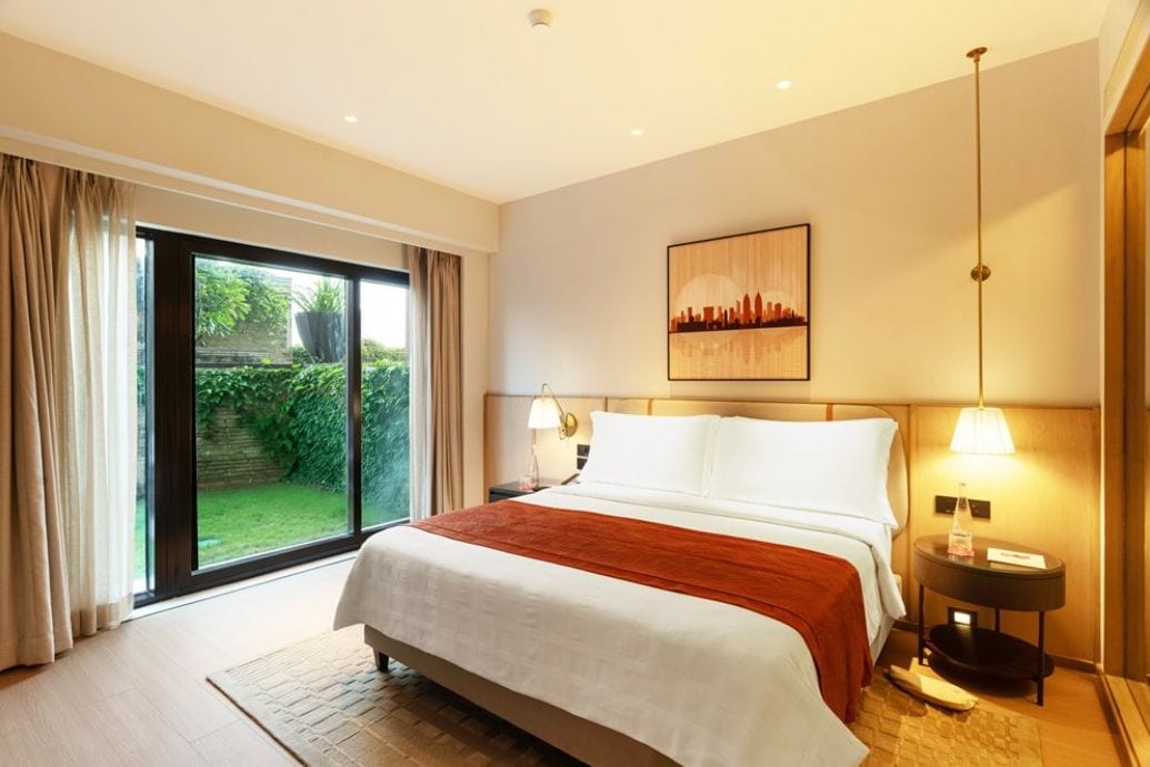 A hotel room with a large bed, a view of greenery through the window, a painting on the wall, and a bedside lamp.