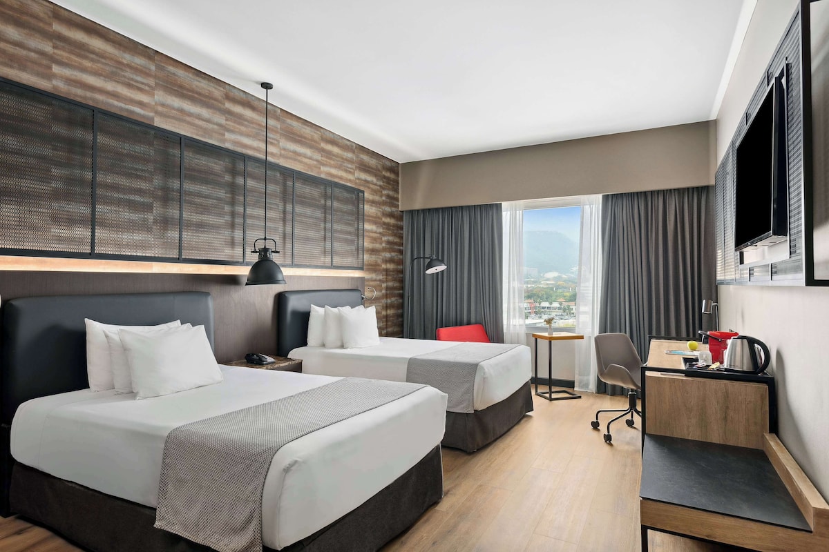 Habitación de hotel con dos camas, un escritorio, una silla, un televisor y vistas a la ciudad.