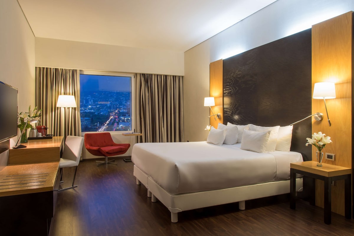 A hotel room with a large bed, a desk, a chair, a TV, and a view of a cityscape through the window.