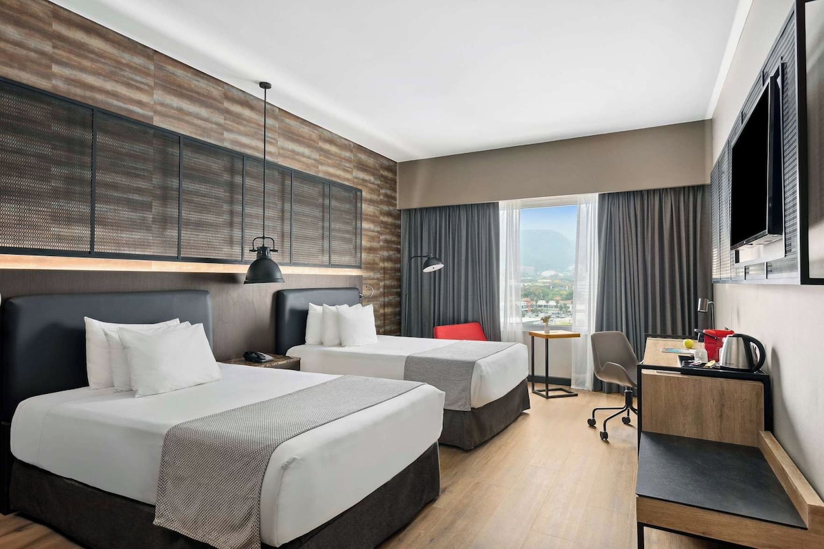 A hotel room with two beds, a desk, a chair, and a large window offering a city view.