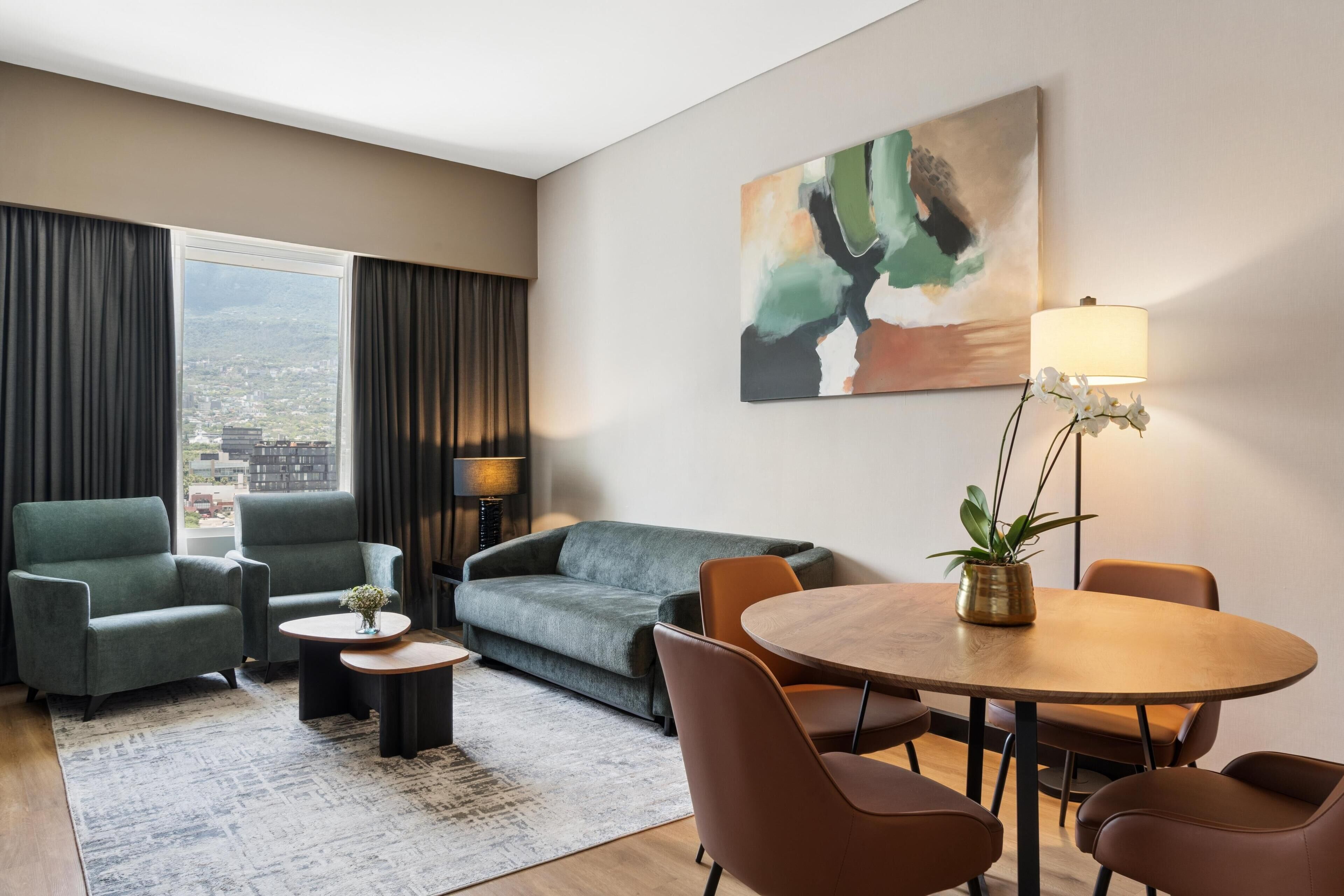 A modern hotel room with a sofa, armchairs, a dining table, a lamp, and a painting on the wall.