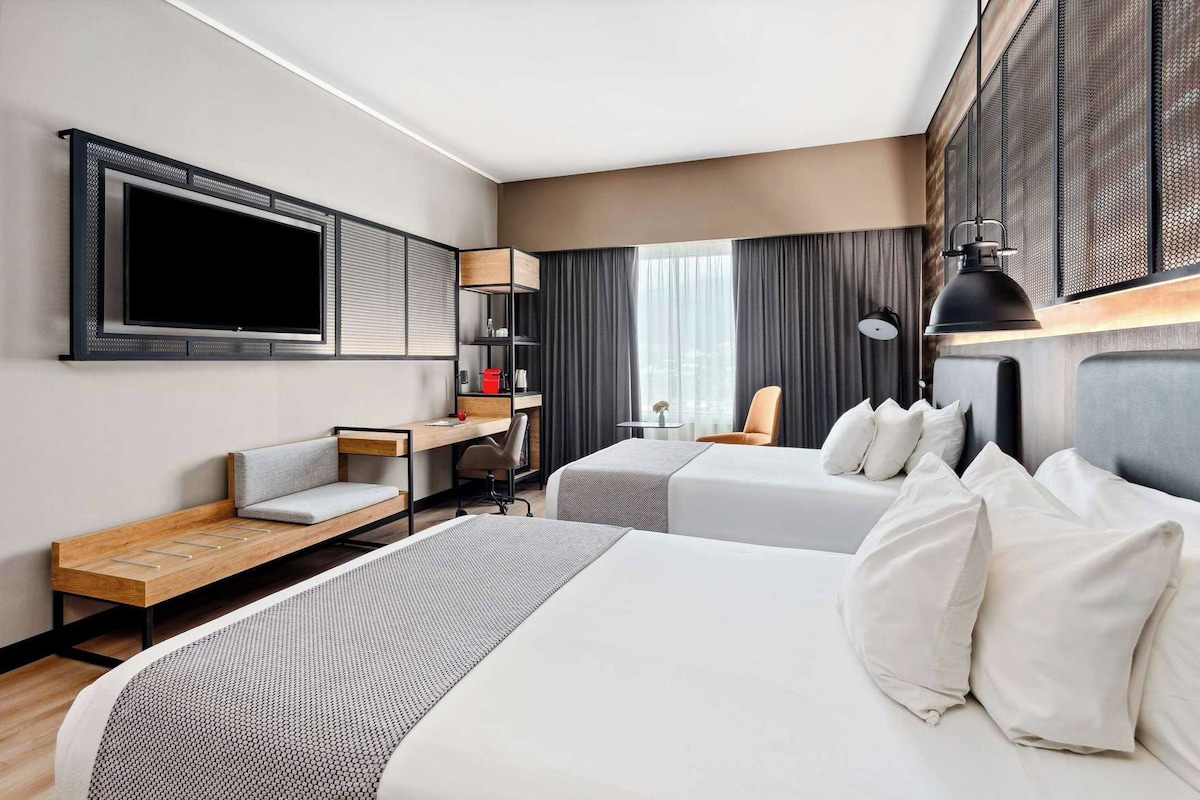 A hotel room with two beds, a desk, a chair, a TV, and a window with curtains.