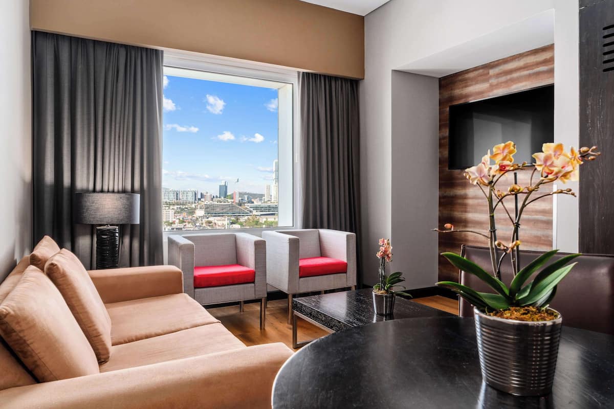 Una habitación de hotel moderna con sofá, dos sillones, una mesa de comedor, una orquídea en maceta y vistas a la ciudad a través de amplias ventanas.