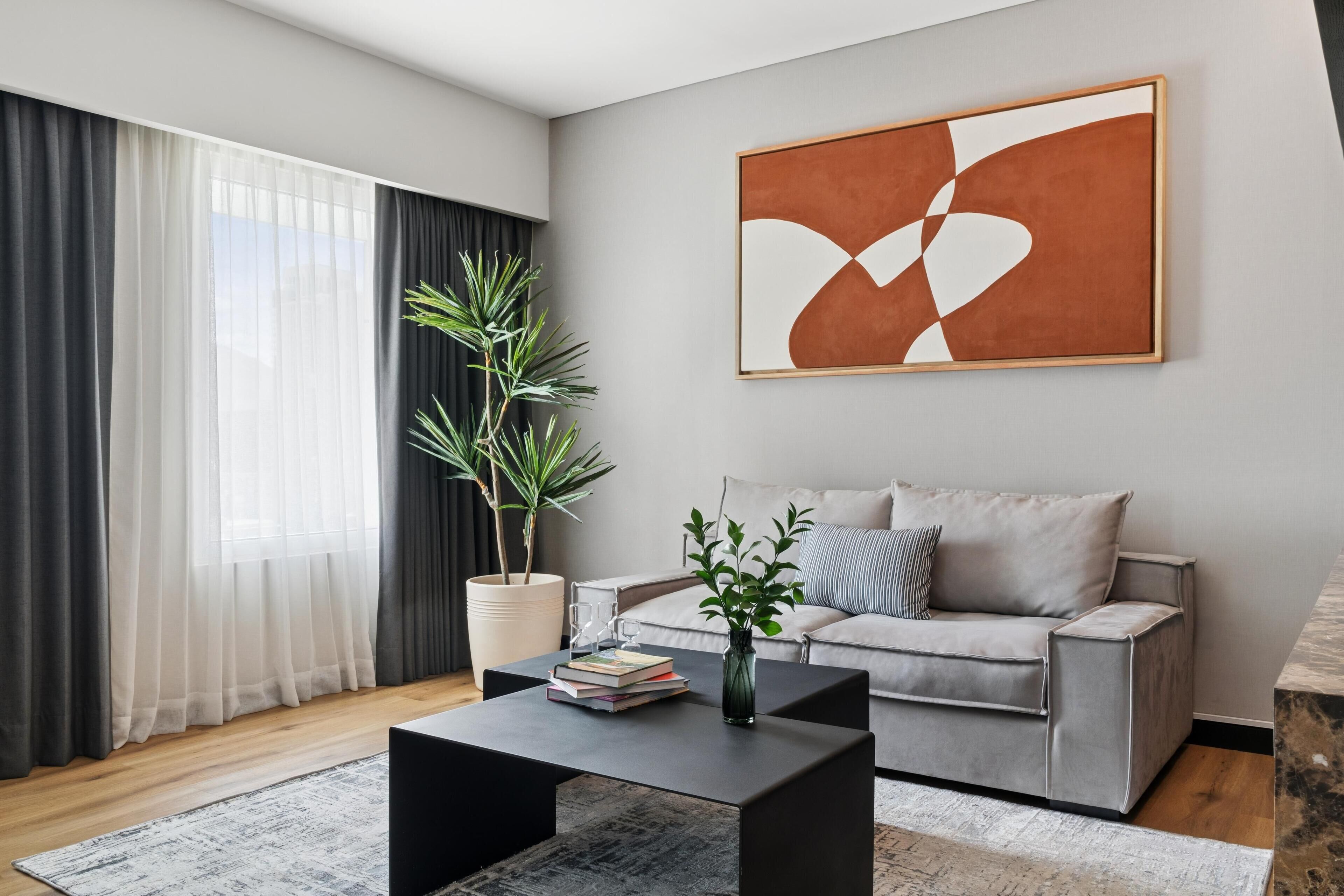 Uma sala de estar moderna com um sofá cinza, uma mesa de centro preta, um grande quadro abstrato, uma planta em vaso e uma janela com cortinas leves.