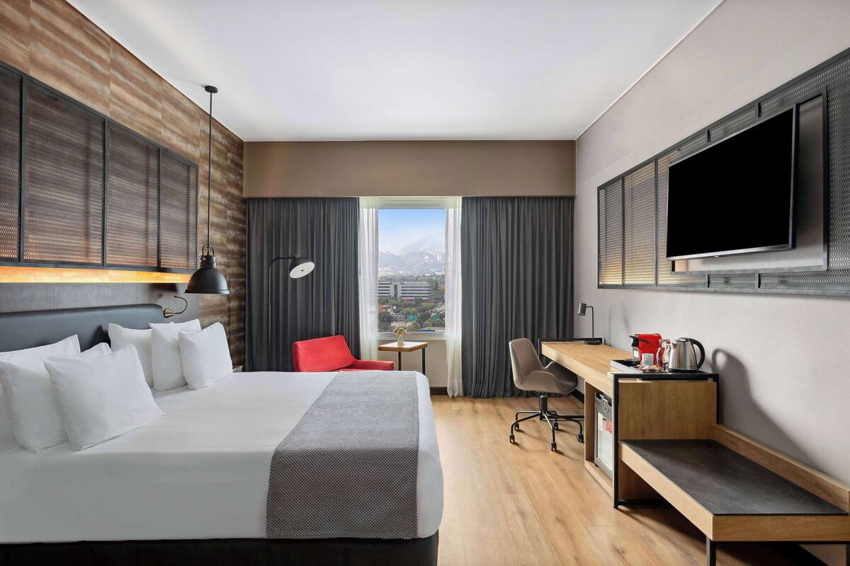 Habitación de hotel con una cama grande, un escritorio, una silla, un televisor y una ventana con vista a la ciudad.
