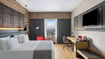 Habitación de hotel con una cama grande, un escritorio, una silla, un televisor y una ventana con vista a la ciudad.