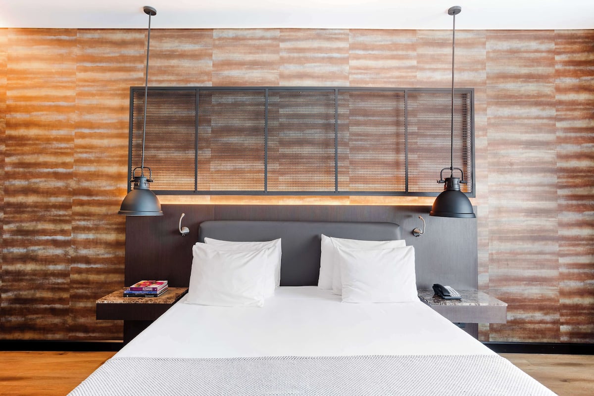 Una habitación de hotel moderna con una cama grande, dos lámparas de noche, una mesita de noche con un libro y un teléfono, y una pared con textura.