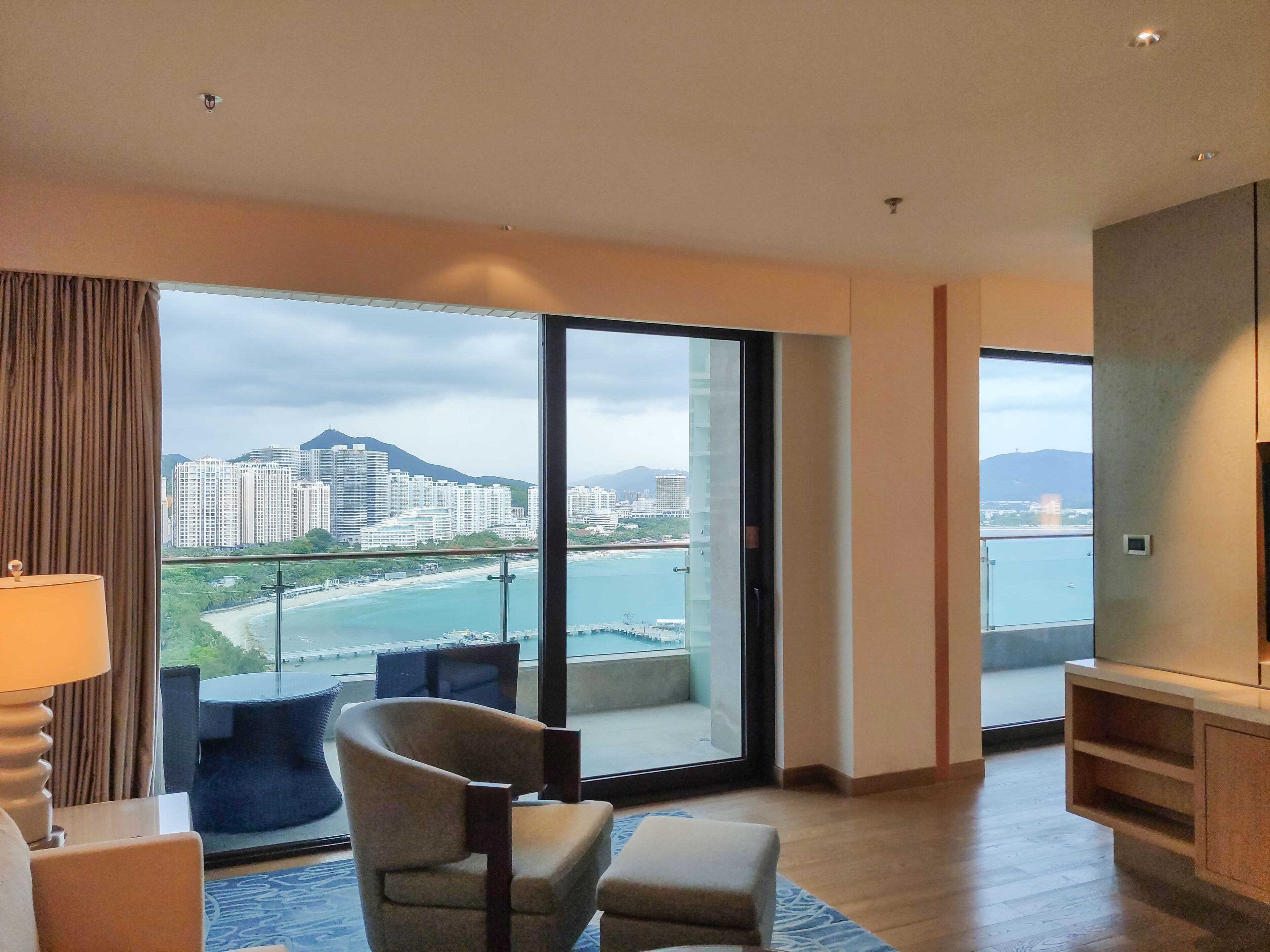 A modern hotel room with a large sliding glass door leading to a balcony, offering a view of a beach and cityscape.