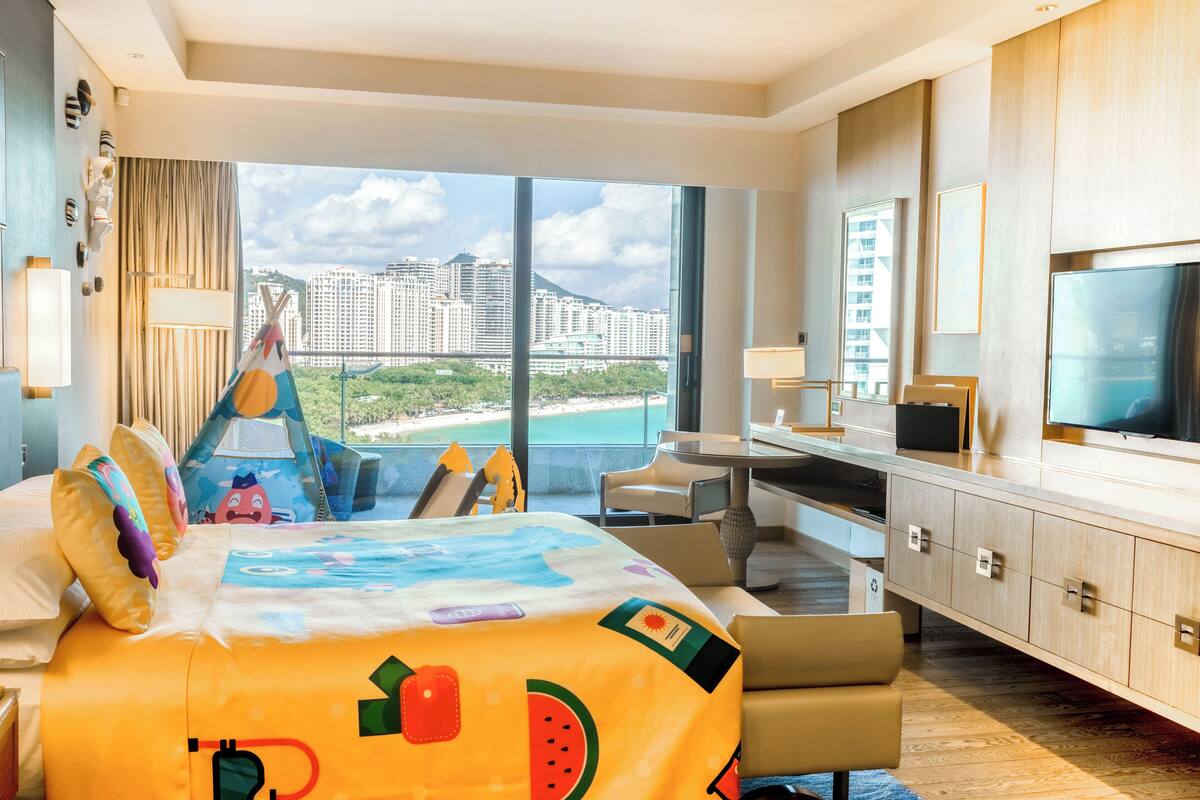 A hotel room with a large bed, a desk, a TV, and a view of a cityscape.