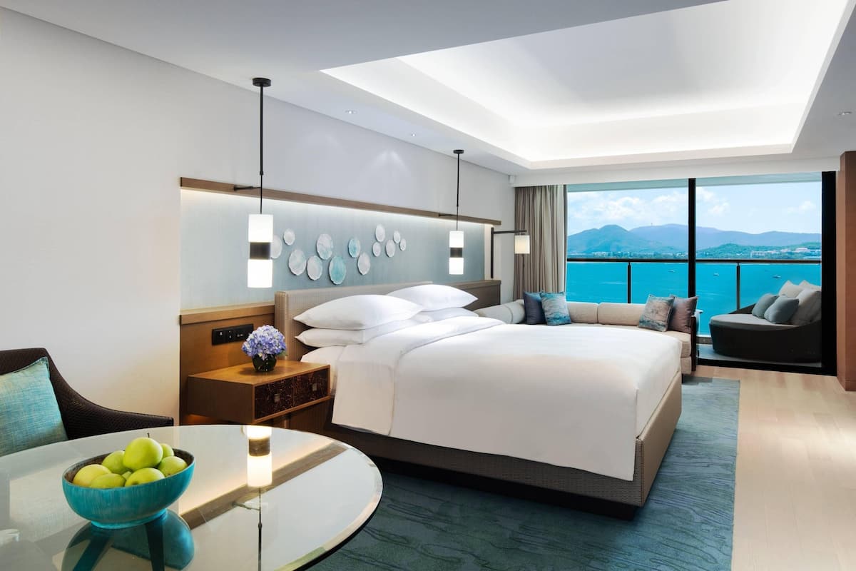 A modern hotel room with a large bed, a dining table, and a view of the sea.