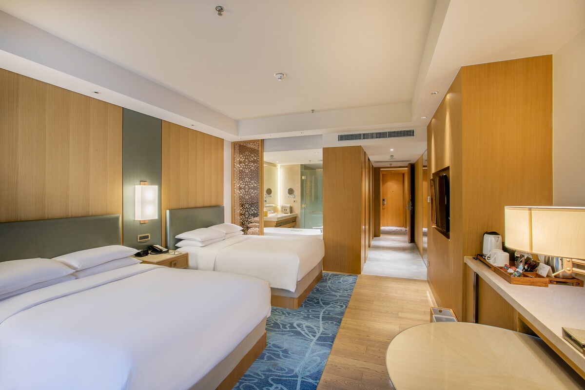 Habitación de hotel con dos camas, un escritorio, un televisor y un baño visible al fondo.